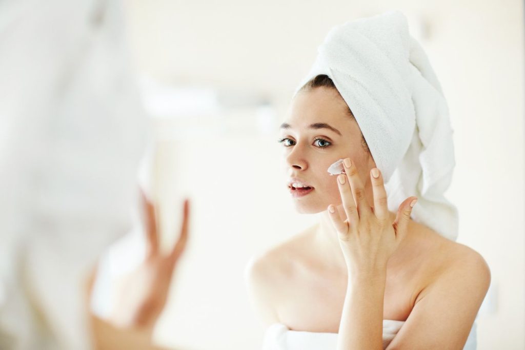 A woman with a towel on her head is putting cream in her face.